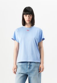 Young person with straight dark hair wearing light blue T-shirt and faded blue jeans, standing against plain white background.