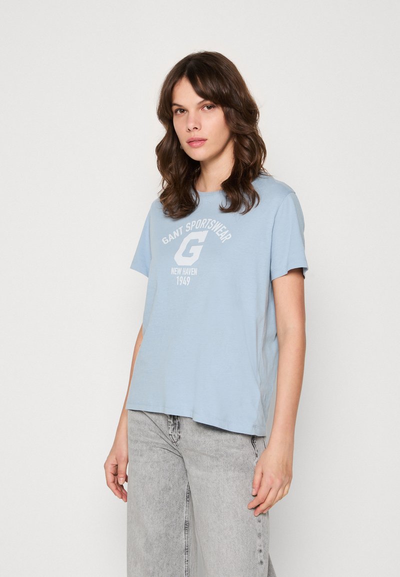 Ung kvinna med vågigt brunt hår, iklädd en ljusblå GANT Sportswear-t-shirt och ljusgrå jeans, stående mot en enfärgad vit bakgrund.