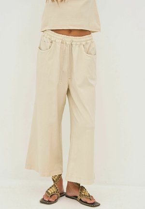 Beige broek met wijde pijpen, elastische tailleband en trekkoord, twee steekzakken aan de voorkant, gedragen met gouden sandalen met studs.