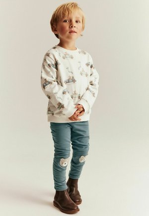 Hvid sweatshirt med dyre- og biltryk, blå leggings med bjørneapplikationer på knæene og brune ankelstøvler.