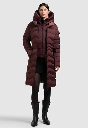 Cappotto imbottito borgogna con cappuccio, caratterizzato da un design trapuntato, zip frontale, tasche laterali e polsini accentuati. Indossato sopra un outfit nero.