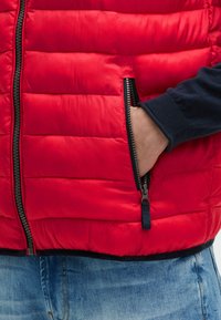 Rote Steppweste mit genoppter Textur und Reißverschlusstaschen. Verfügt über einen schwarzen Reißverschluss und ein dunkelblaues Bündchendesign. Blaue Jeans darunter sichtbar.
