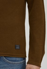 Brown knitted sweater with a textured pattern, featuring a black label with white text. The sweater has a ribbed hem and cuffs.
