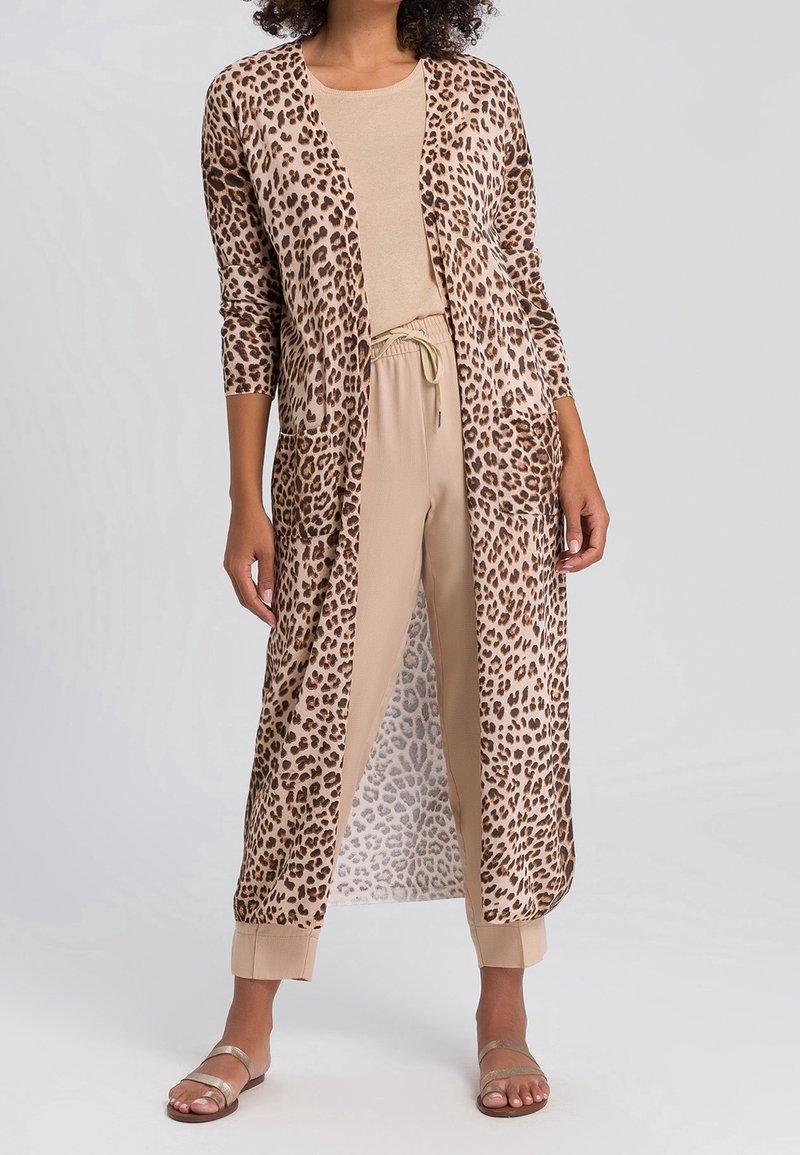 Femme portant un long cardigan ouvert imprimé léopard sur un top beige et un pantalon avec des revers roulés, ainsi que des sandales dorées, debout sur un fond clair.