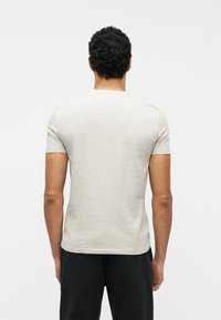 Άνδρας με σγουρά σκούρα μαλλιά, φοράει ανοιχτό γκρι κοντομάνικη μπλούζα και μαύρο παντελόνι, στέκεται με την πλάτη προς το μέρος μας πάνω σε λευκό φόντο.