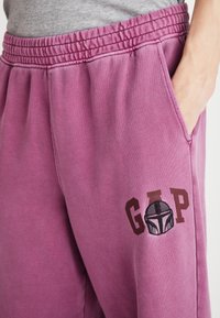 Flis hlače v utišanem roza odtenku z elastičnim pasom, stranskimi žepi in vezenim logotipom "GAP" ob risbi čelade.