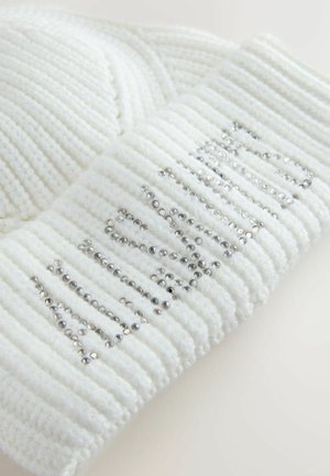 Bela pletena zimska kapa s rebrastom teksturom, koja na preklopu ima riječ "zima" ukrašenu srebrnim šljokicama.