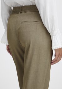 Khaki nohavice na mieru s hladkou textúrou, s jednou zadnou vreckom a pútkami na opasok, v kombinácii s bielou blúzkou s volánovými rukávmi.