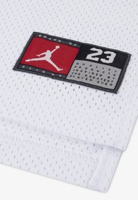 Λευκό ύφασμα από δίχτυ με ραμμένη μαύρη και κόκκινη ετικέτα που αναγράφει «Brand of 23 Flight» και μια σιλουέτα παίκτη. Ξεχωρίζει για την ελαφριά υφή του.