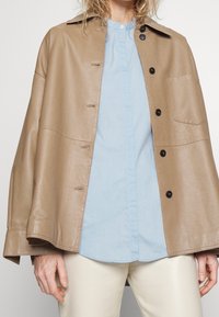 Brun läderjacka med lös passform, som har en krage, framknäppning och en enda ficka. Underjackan syns en ljusblå skjorta med knappar.