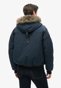 Marineblå puffjakke med en hætte beklædt med pels, ribbede manchetter og en indsnævret kant, med en læderstrop detalje bagpå.