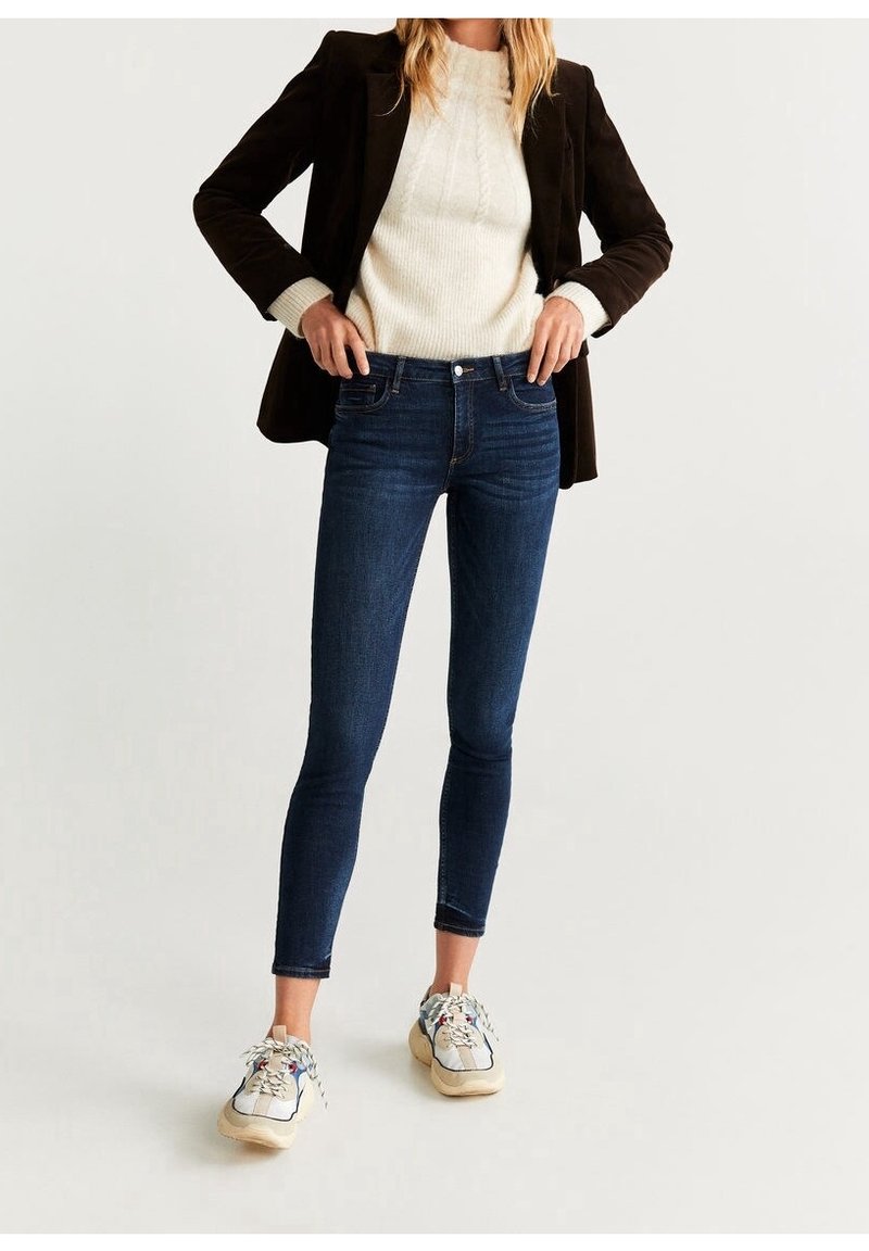 Femme portant un blazer marron foncé, un pull en maille crème, un jean skinny bleu foncé et des baskets beiges, debout, les mains sur les hanches.