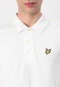 Vit polo t-shirt med texturerad tyg, utrustad med en broderad logotyp i gult och svart. Tre-knapps placket och kontrasterande krage.