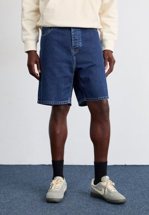Mørkeblå denimshorts i knælængde med lige skæring. Har lynlås foran og traditionelt fem-lomme design. Båret med grå sneakers.