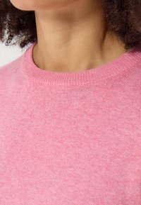 Růžový pletený svetr s kulatým výstřihem. Tkanina vypadá měkce s jemnou texturou a rovnoměrnou barvou po celém povrchu.