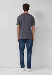 Mann in einem schlichten grauen T-Shirt mit kurzen Ärmeln, blauen Jeans und weißen Turnschuhen, der einer hellgrauen Wand gegenübersteht.