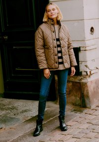 Tan quiltad jacka med tryckknappar, randig tröja, blå jeans och svarta ankelboots. Modellen står på kullersten nära en svart dörr.