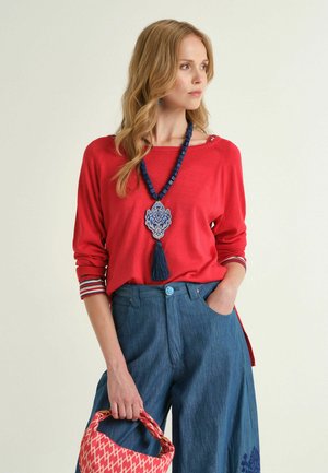 Donna con maglia rossa a maniche lunghe, pantaloni larghi blu, che tiene una borsa rossa con motivi, indossa una lunga collana blu con perline e nappina, guarda di lato.
