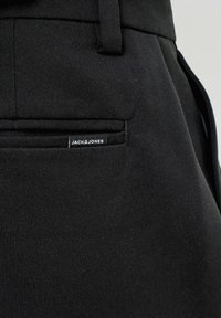 Gros plan sur un pantalon en tissu noir avec une poche arrière et une petite étiquette de marque Jack & Jones cousue près de la couture de la poche.