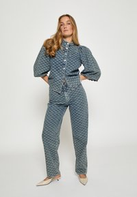 Denim obleka, ki vključuje jakno s ovratnikom in gumbi z napihnjenimi rokavi ter usklajenimi hlačami z visokim pasom, oboje v modrem vzorcu, dopolnjena s bež petkami.