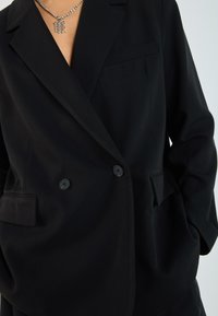 Black blazer made of smooth fabric with a structured fit. Features a chest pocket, two front pockets, and two black buttons.