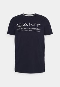 T-paita tummansinisestä puuvillasta, jossa on lyhyet hihat ja valkoisella painettuna teksti "GANT AMERICAN SPORTSWEAR SINCE 1949" rinnassa.