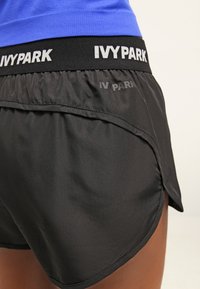 Primo piano di pantaloni sportivi neri Ivy Park con vita elastica e logo bianco, indossati con una maglia blu.