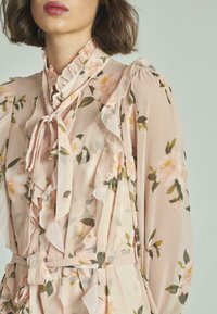 Blusa floral en suave tela rosa con detalles de volantes, cuello con lazo y botones. Presenta hojas verdes y un patrón floral en toda la prenda.