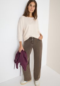 Beiger Strickpulli, braune weite Hose und weiße Sneaker mit blauen Akzenten; das Modell hält einen kastanienbraunen Kapuzenpullover. Glatte Texturen, lässiges Design.
