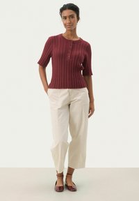 Pull en maille bordeaux à manches courtes avec détail de bouton, associé à un pantalon large crème et des ballerines bordeaux. Tissu texturé sur l'ensemble.