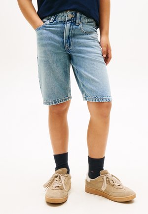 Kind draagt lichtblauwe denim shorts, zwarte sokken, beige suède sneakers en een donkerblauw shirt, staat met één hand in de zak.