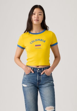 Kobieta z długimi ciemnymi włosami ubrana w żółty krótki t-shirt z napisem „Colombia” i flagą, zestawiony z niebieskimi podartymi dżinsami, ręce w kieszeniach.