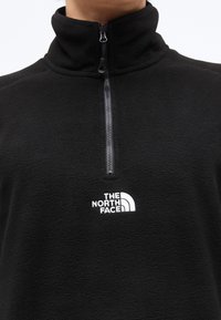 Μαύρο φλις πουλοβεράκι με φερμουάρ στη μέση του μπροστινού μέρους και ψηλό γιακά, με λευκό λογότυπο "The North Face" στο στήθος.