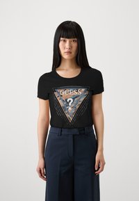 T-shirt noir avec un graphique en forme de triangle comportant le texte "GUESS", entouré d'accents en points. Le t-shirt a un col rond et des manches courtes.