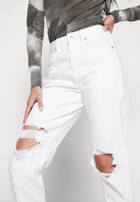 Bílé roztrhané džíny ve střihu rovného nohavic s roztrhanými kolenními záplaty. Kombinované s texturovaným šedým topem s dlouhým rukávem.