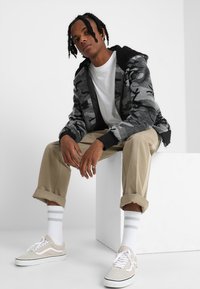 Jachetă bomber cu camuflaj gri și accente negre, tricou alb, pantaloni khaki și adidași bej. Modelul este așezat pe o platformă albă.
