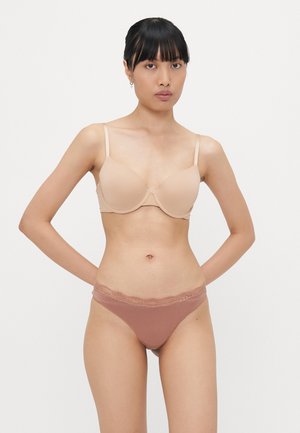 Beige gevoerde bh met dunne bandjes, gecombineerd met bruine string met kantaccenten. Glad materiaal, naadloos ontwerp. Model staat met handen op de heupen.