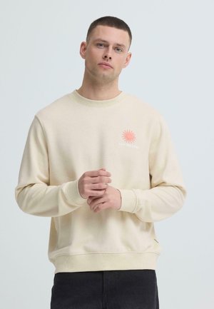 Młody mężczyzna z krótkimi włosami, ubrany w kremową bluzę z małym czerwonym logo słońca i napisem „Sun Seeker Fest” na piersi, z rękami złączonymi.