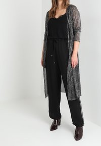 Gennemsigtig sort duster med sølvmetaltrådsdetaljer, båret over en sort tanktop og sorte bukser med sidelommer, afsluttet med burgunderrøde hæle.