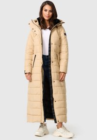 Manteau long beige en duvet avec texture matelassée, doublure intérieure noire, fermeture éclair noire et pressions ; deux poches latérales et une capuche. Porté sur un jean et des baskets.