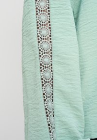 Υφασμα ανοιχτού πράσινου χρώματος με υφή στην επιφάνεια που διαθέτει κάθετη ταινία περίτεχνων κυκλικών δαντελένιων λεπτομερειών.