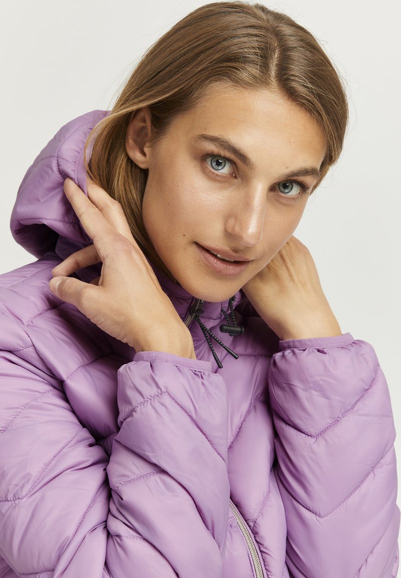 Chaqueta de peluche lavanda con capucha, que presenta un diseño acolchado, puños elásticos y un cierre frontal con cremallera. Textura suave y brillante.