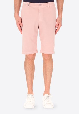 Uomo con pantaloncini su misura rosa chiaro e sneaker bianche, in piedi con le mani rilassate lungo i fianchi davanti a uno sfondo semplice.