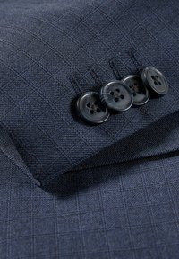 Marineblauwe stof met een subtiel ruitpatroon, voorzien van een manchet met vier donkere knopen, wat textuur en detail aan het ontwerp toevoegt.