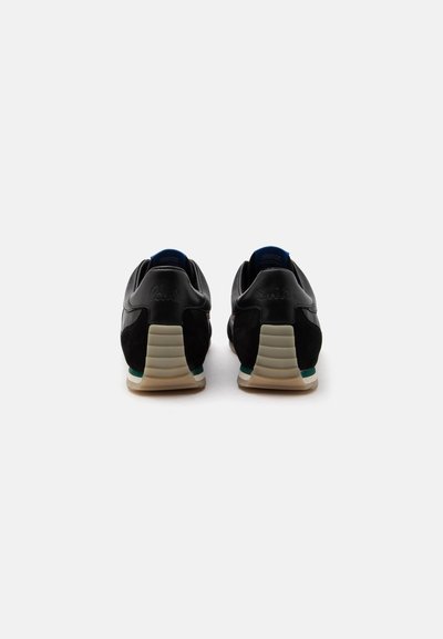 Paire de baskets basses noires vues de dos, avec des semelles beige et des accents verts et blancs subtils sur fond blanc.