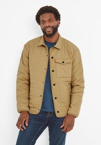 Tan quilted jacket with a front pocket, black buttons, and a blue shirt underneath. The fabric is smooth with a padded texture.