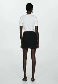 Mujer con cabello corto que lleva una blusa blanca de mangas cortas, una mini falda negra texturada, pendientes de aro y sandalias negras de tacón, de pie de espaldas.