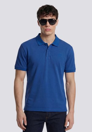 Joven con gafas de sol negras, una camiseta polo azul de manga corta y vaqueros oscuros, de pie frente a un fondo claro y liso.