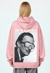 Oversized roze hoodie met een vintage zwart-wit portretprint en de tekst "ART IS EXPENSIVE" op de achterkant. Zacht materiaal.