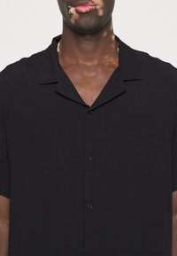 Camicia nera a maniche corte con colletto morbido, caratterizzata da una chiusura frontale con bottoni. Texture liscia e design minimalista.